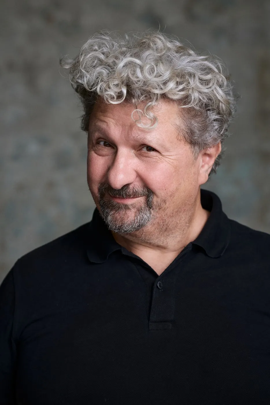Porträt von Aykut Kayacık, schwarzes Polo vor grauem Studiohintergrund, verschmitztes Lächeln